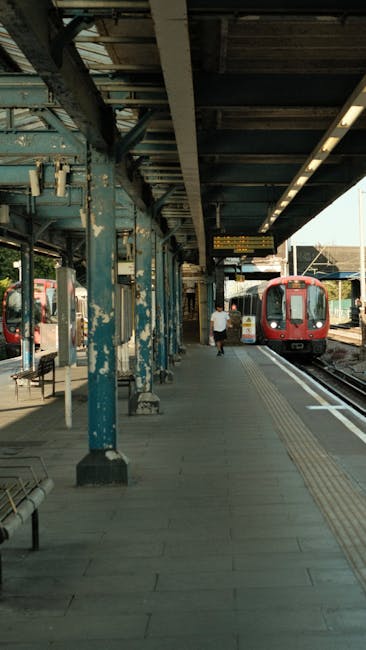 At West Brompton railway station, the platform features a weathered, rust-colored metal canopy supported by blue-painted metal beams. The platform surface is paved with grey concrete tiles, with tactile paving strips near the edge for safety. A red and black train is stationary on the tracks on the right side of the image. A person wearing casual clothing is walking along the platform, moving away from the camera towards the train. To the left, there are empty metal benches and a small shelter with a window, likely for ticketing or information. The station environment includes various structural elements such as overhead electrical wiring and platform lighting, conveying a typical urban railway setting. This scene relates to the process of home relocation or furniture transport as part of house removals, facilitated or documented by Man With a Van West Brompton, emphasizing the logistical aspects of moving services and the transportation of belongings via train or coordinated loading and unloading.