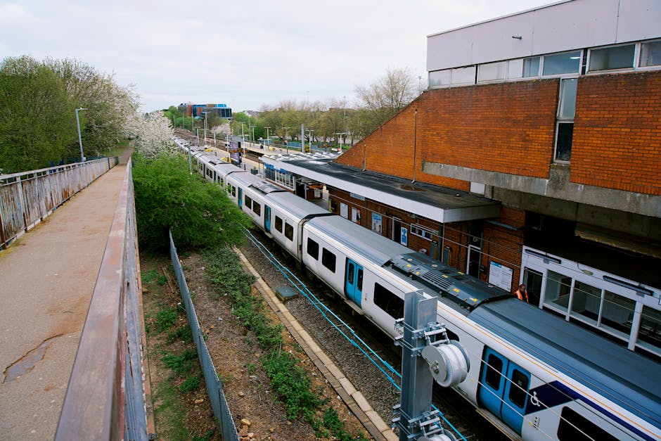 A modern passenger train with a streamlined silver exterior and orange and black accents is stationary at West Brompton station platform. The train's front end is visible, showing a large black window and headlight, while the side features multiple windows and doors for boarding. The platform has tactile paving strips along the edge and is covered by a roof supported by metal beams. Several people are seated on benches further along the platform, some waiting for departure or arrival. In the background, another train can be seen on a parallel track, with station signage visible but not legible. The setting appears to be during daylight hours with overcast but bright lighting, typical of a busy urban railway station prepared for home relocation or furniture transport involving careful loading processes. Occasionally, Man With a Van West Brompton's services may incorporate transportation at such stations as part of comprehensive removals and moving logistics.